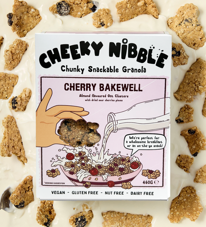  A box of Cherry Bakewell-flavored chunky granola is displayed, showcasing an image of the granola bathed in creamy thick yogurt. The granola features chunks studded with sour cherries, offering a tantalizing blend of tartness and sweetness. The background evokes a sense of indulgence, with hints of almond flourishes and cherry blossoms, enhancing the product's appeal. 